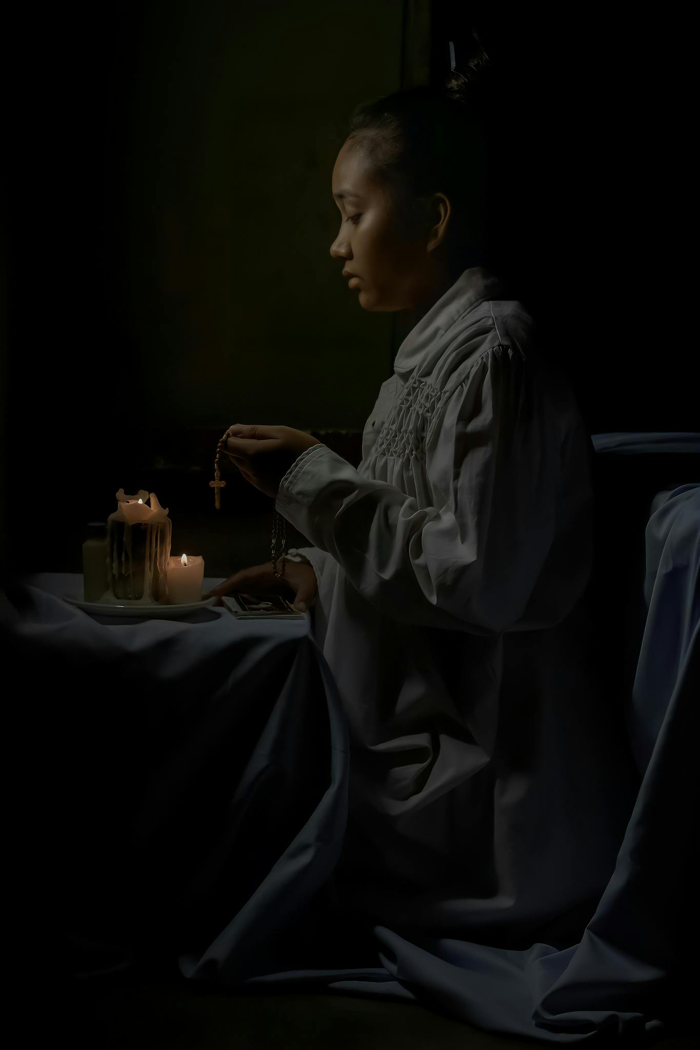 Side view of African American female teen in religious clothes with cross praying against flaming candles on black background